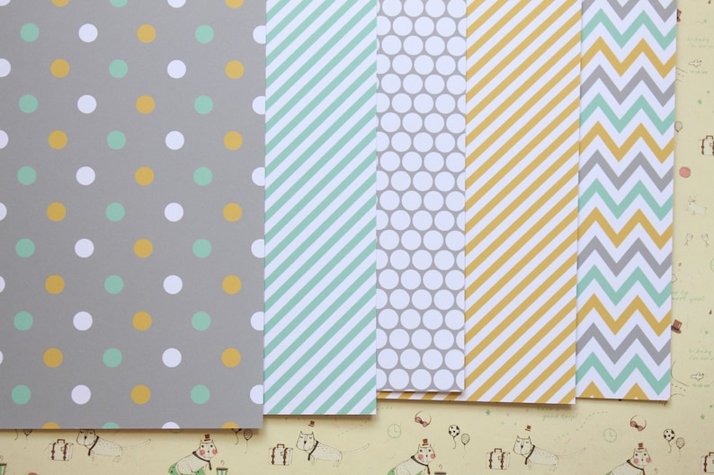 May include: Five sheets of patterned paper with different designs. The first sheet has a gray background with white and yellow polka dots. The second sheet has a light blue and white striped pattern. The third sheet has a gray background with white polka dots. The fourth sheet has a yellow background with white stripes. The fifth sheet has a gray and light blue chevron pattern.