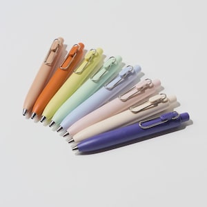 May include: A collection of pastel-coloured pens in shades of peach, orange, yellow, mint green, light blue, pink, cream, and purple. Each pen features a silver clip and a black ink tip. The pens are arranged in a fan shape on a white surface.