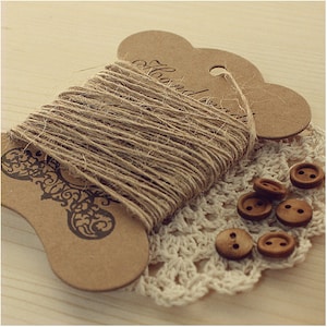 May include: A spool of natural twine wrapped around a brown cardboard tag with the text "Hand Made" printed on it. The twine is sitting on a white crocheted doily with brown buttons scattered around it.