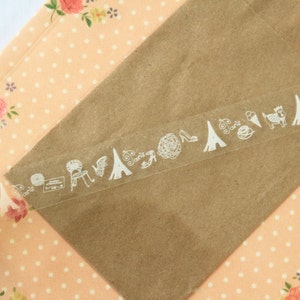 May include: A roll of clear washi tape with white decorative designs, including a chair, a house, a heart, a shoe, and a Eiffel Tower. The tape is on a peach-colored background with white polka dots and floral patterns.