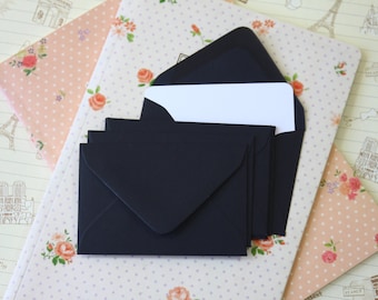 May include: A set of black envelopes with a white card inside. The envelopes are on a pink and white patterned surface.