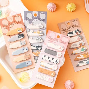 May include: Multiple packs of "The Cat Note" index stickers, each with 180 pieces. The stickers showcase cat-themed designs, such as cat shapes and paw prints in diverse colours. The background is a light orange.
