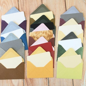 以下が含まれることがあります： 木製の表面に並んだ、さまざまな質感と模様のカラフルな封筒の束。