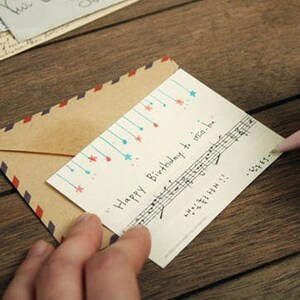 May include: A white card with blue and red stars and musical notes. The card says "Happy Birthday to win-win-love". The card is being held by a hand and is partially covered by a brown envelope with a red and blue striped border.