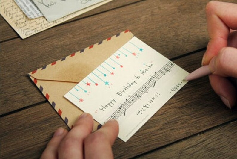 May include: A white card with blue and red stars and musical notes. The card says "Happy Birthday to win-win-love". The card is being held by a hand and is partially covered by a brown envelope with a red and blue striped border.