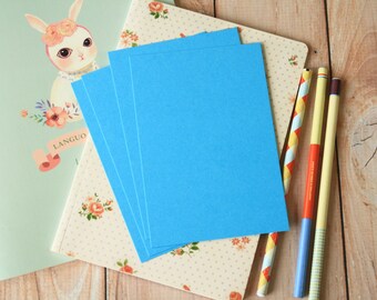 May include: A stack of five blue cardstock sheets on a patterned paper background with three colorful pencils. The paper has a floral pattern with white polka dots. The pencils are yellow, red, and blue with white stripes.