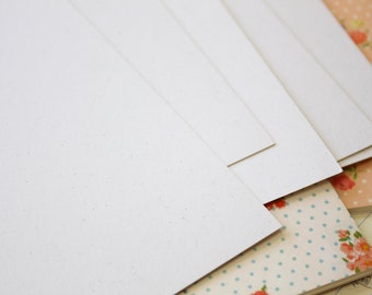 May include: Stack of white paper sheets with a speckled texture, arranged on top of a patterned fabric with white polka dots and pink flowers.