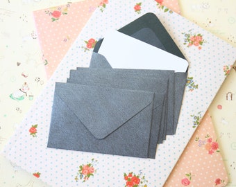 May include: A stack of six gray envelopes with a white card inside the top envelope. The envelopes are on a patterned surface with pink and white flowers.