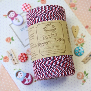 May include: A spool of red, white, and navy blue baker's twine with a kraft paper label that reads "Beautiful Baker's Twine." The twine is approximately 100 meters long. The background includes decorative buttons, clothespins, and a floral patterned paper.