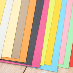 May include: A collection of 15 different colored cardstock paper sheets arranged in a fan shape. The colors include yellow, cream, beige, brown, orange, black, pink, green, light blue, dark blue, and red.