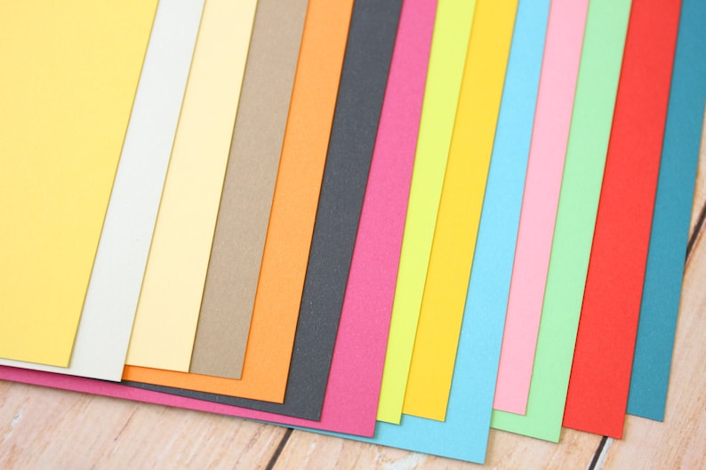 May include: A collection of 15 different colored cardstock paper sheets arranged in a fan shape. The colors include yellow, cream, beige, brown, orange, black, pink, green, light blue, dark blue, and red.