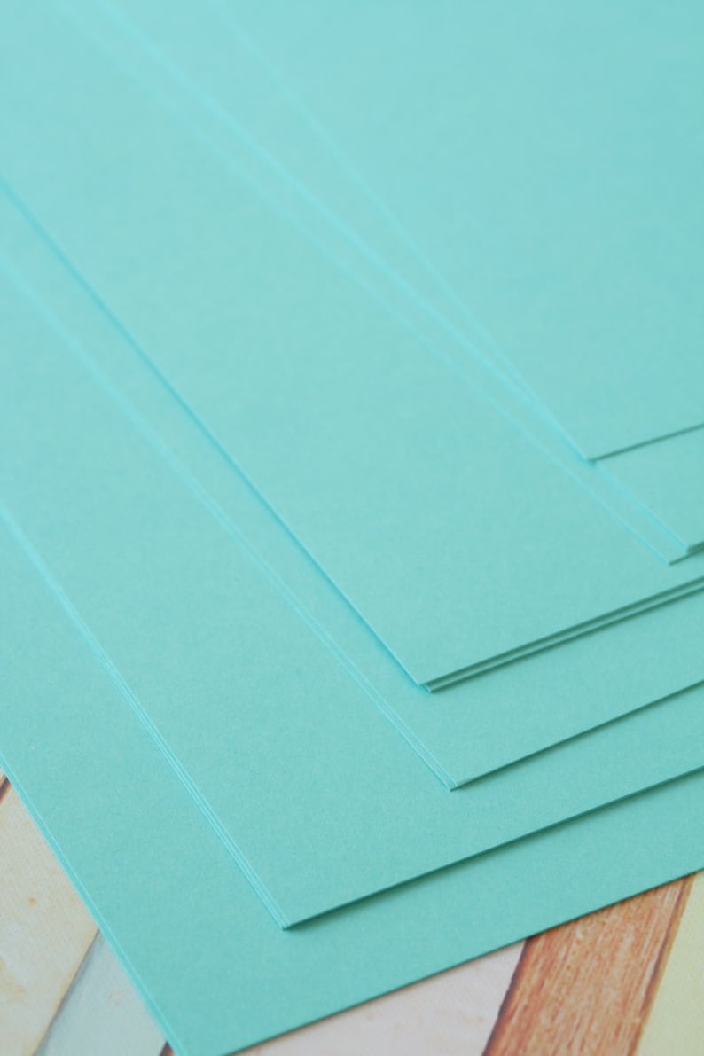 K&ouml;nnte beinhalten: Ein Stapel hellt&uuml;rkisfarbener Papierb&ouml;gen. Das Papier hat eine glatte Textur und ist diagonal angeordnet, wobei die Kanten der B&ouml;gen sichtbar sind. Der Hintergrund ist eine Mischung aus Holz und hellen Farben.