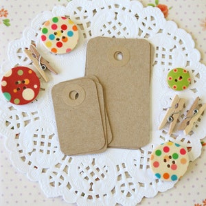 May include: A set of two brown kraft paper gift tags with a hole punched in the top. The tags are surrounded by colourful polka dot buttons and wooden clothes pegs on a white doily.
