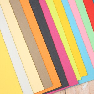 May include: A collection of colorful paper sheets arranged in a diagonal pattern. The colors include yellow, white, light yellow, brown, orange, black, pink, green, light blue, blue, and red.
