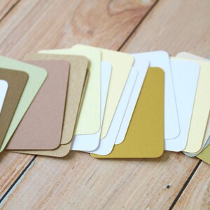 May include: A collection of rectangular paper cards with rounded corners, arranged in a fan-like overlapping pattern. The cards feature a variety of solid colors including white, cream, yellow, mustard, tan, brown, light green, and sage green.