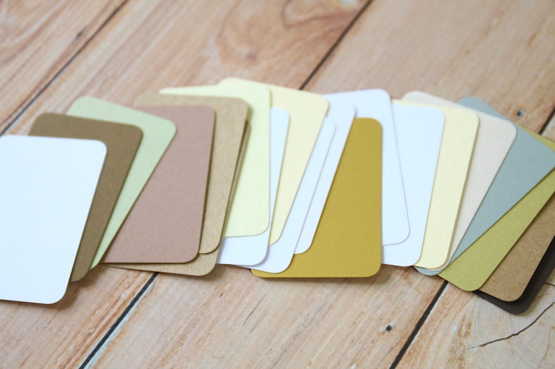 May include: A collection of rectangular paper cards with rounded corners, arranged in a fan-like overlapping pattern. The cards feature a variety of solid colors including white, cream, yellow, mustard, tan, brown, light green, and sage green.