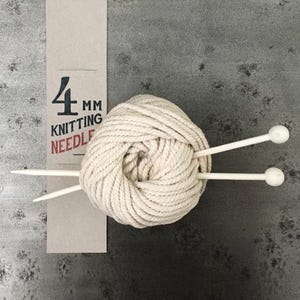 May include: A ball of cream-colored yarn with two white knitting needles. A cardboard label displays "4 mm Knitting Needle". The yarn is tightly wound, and the needles are inserted through the center of the ball. The background is a mottled grey.