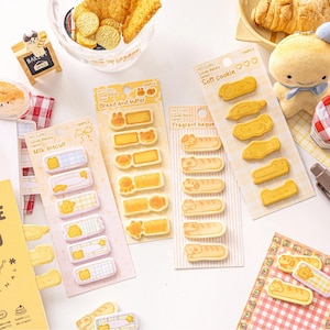 May include: A selection of stationery items with a bakery theme. The image shows sticker sheets with bread, biscuit, and cookie designs, alongside a plush toy and small decorative items. The stickers are in various shapes and colours.