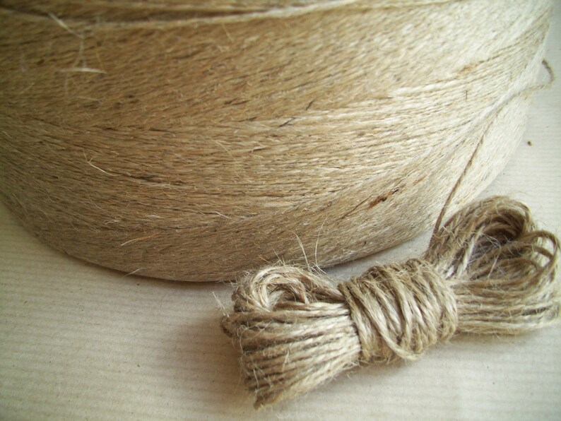 May include: A roll of natural twine, made from jute fiber, is shown with a loose end of the twine. The twine is a light brown color and has a rough texture.