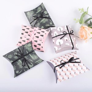 May include: Five pillow boxes with different patterns. The boxes are made of cardboard and have a black ribbon tied around them. The patterns include trees, leaves, and geometric shapes. The boxes are all in shades of pink, green, and black.