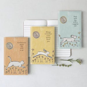 May include: Three small notebooks with animal illustrations. One features a cat, another a bunny, and the third a dog. Each notebook has a different colour and a quote. The open notebook has lined pages.