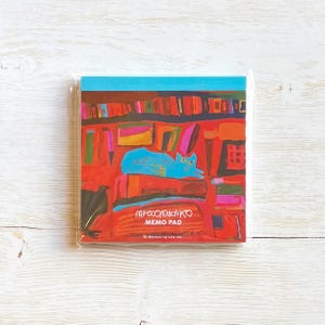 May include: A square memo pad featuring a vibrant illustration of a blue cat resting on books. The artwork includes a bookshelf and a black bird. The text "mirocomachiko MEMO PAD" is displayed on the pad.