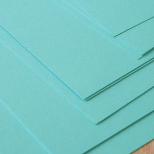 K&ouml;nnte beinhalten: Ein Stapel rechteckiger, hellt&uuml;rkisfarbener Papierb&ouml;gen. Das Papier ist diagonal angeordnet, wobei die Kanten der B&ouml;gen sichtbar sind. Das Papier ist glatt und scheint neu zu sein.