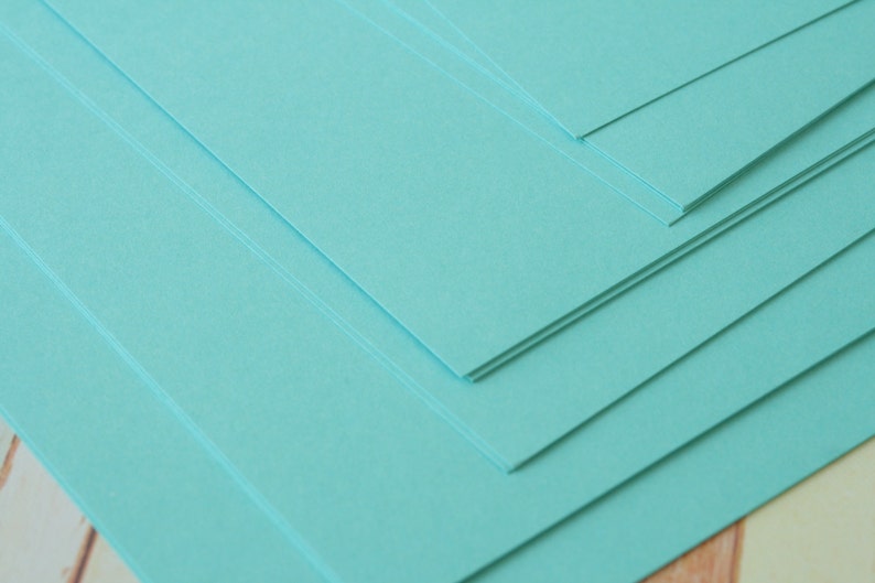 K&ouml;nnte beinhalten: Ein Stapel rechteckiger, hellt&uuml;rkisfarbener Papierb&ouml;gen. Das Papier ist diagonal angeordnet, wobei die Kanten der B&ouml;gen sichtbar sind. Das Papier ist glatt und scheint neu zu sein.
