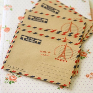 May include: A stack of brown paper envelopes with red and blue stripes. The envelopes have a vintage Parisian theme with the text "Par Avion" and "Paris-VII 14 Juil 67" printed on them.
