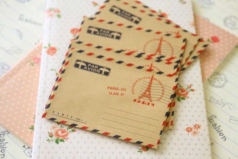 May include: A stack of brown paper envelopes with red and blue stripes. The envelopes have a vintage Parisian theme with the text "Par Avion" and "Paris-VII 14 Juil 67" printed on them.