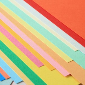 Może przedstawiać: Stos kolorowych arkuszy papieru w r&oacute;żnych odcieniach niebieskiego, zielonego, ż&oacute;łtego, pomarańczowego, r&oacute;żowego i czerwonego. Papier jest ułożony po przekątnej, tworząc żywy i wizualnie atrakcyjny wyświetlacz.