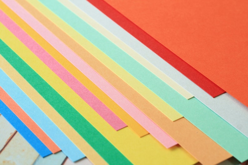 Może przedstawiać: Stos kolorowych arkuszy papieru w r&oacute;żnych odcieniach niebieskiego, zielonego, ż&oacute;łtego, pomarańczowego, r&oacute;żowego i czerwonego. Papier jest ułożony po przekątnej, tworząc żywy i wizualnie atrakcyjny wyświetlacz.