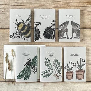 May include: A selection of small notebooks, each with a different nature-inspired illustration and a short quote. The designs include a bee, badgers, hares, a dragonfly, oak leaves, and potted plants. The text includes phrases such as "Bee happy" and "Dream your dreams."