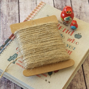 May include: A roll of natural twine wrapped around a cardboard spool with a hole in the centre. The spool is sitting on top of a book with a white cover and colourful patterned edges. The book has the text "A friend is someone who..." printed on the cover. Two small wooden dolls are sitting on the book.