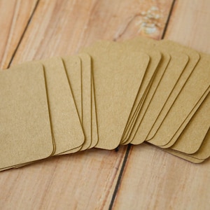 May include: A stack of rectangular kraft paper tags with rounded corners. The tags are light brown in color and feature a smooth, matte surface. Each tag is uniform in size and shape, arranged in a slightly fanned-out pile to show the edges.