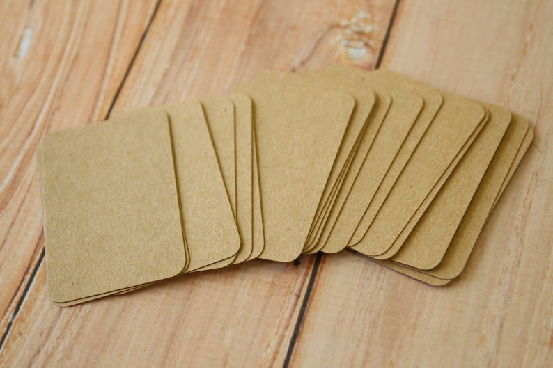 May include: A stack of rectangular kraft paper tags with rounded corners. The tags are light brown in color and feature a smooth, matte surface. Each tag is uniform in size and shape, arranged in a slightly fanned-out pile to show the edges.
