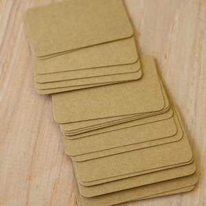 May include: A stack of rectangular, light brown kraft paper cards with rounded corners. The cards are arranged in a slightly fanned, overlapping pile, displaying a smooth, matte texture and a consistent, neutral tan color throughout the set.