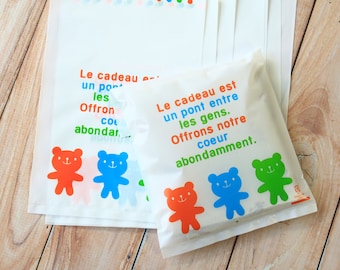 May include: White gift bags with colorful teddy bear illustrations and the text "Le cadeau est un pont entre les gens. Offrons notre coeur abondamment."
