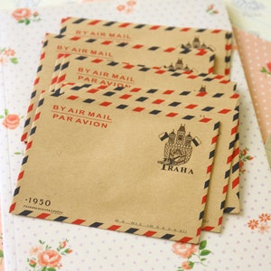 May include: A stack of brown envelopes with red and blue stripes. The envelopes have the text "BY AIR MAIL PAR AVION" and a small image of a building with the word "TRAHA" printed on them. The envelopes are on a white background with a floral pattern.