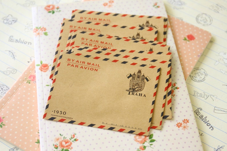 May include: A stack of brown envelopes with red and blue stripes. The envelopes have the text "BY AIR MAIL PAR AVION" and a small image of a building with the word "TRAHA" printed on them. The envelopes are on a white background with a floral pattern.