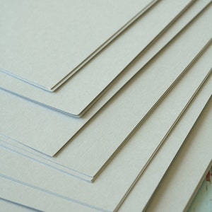 Może przedstawiać: Stos jasnoszarego papieru kartonowego. Papier jest gładki i ma subtelną fakturę.