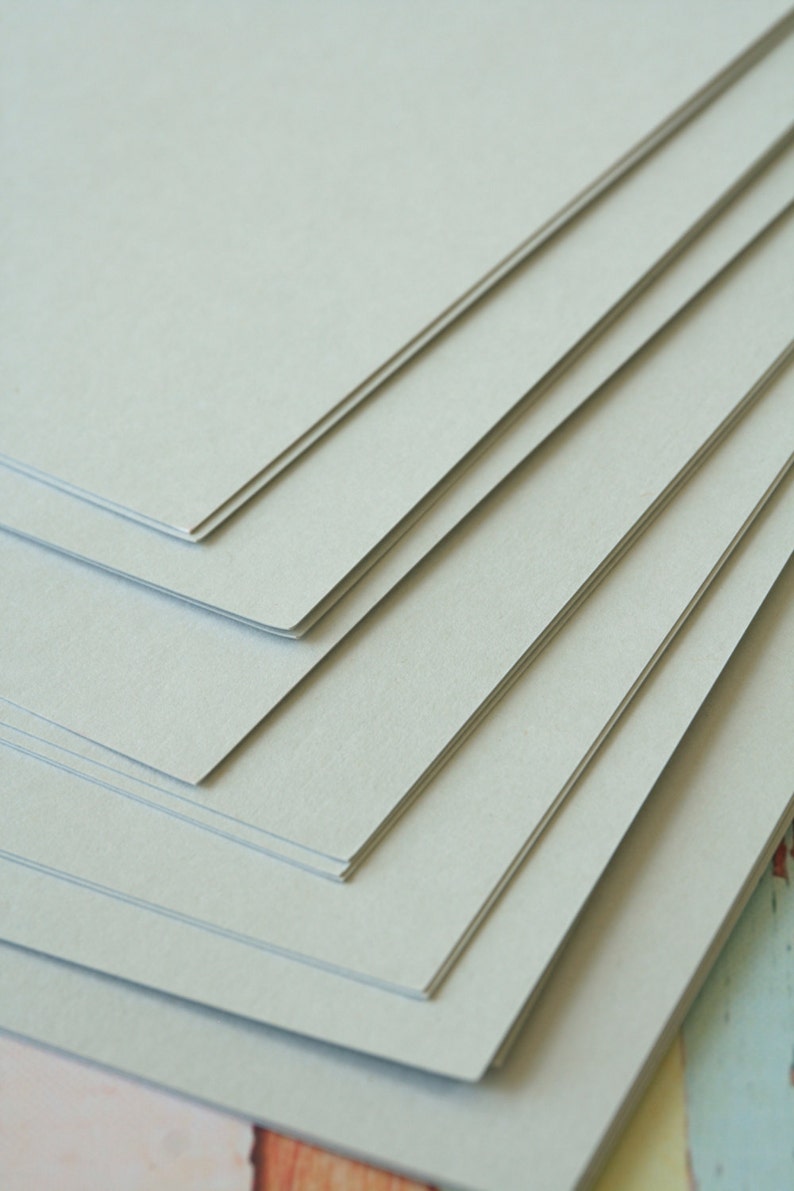 Może przedstawiać: Stos jasnoszarego papieru kartonowego. Papier jest gładki i ma subtelną fakturę.