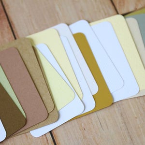 May include: A collection of rectangular cardstock samples arranged in a fan shape. The set features a variety of colors including white, cream, light yellow, mustard yellow, sage green, tan, and dark brown, with some cards showing a textured finish.
