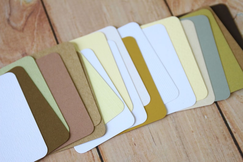 May include: A collection of rectangular cardstock samples arranged in a fan shape. The set features a variety of colors including white, cream, light yellow, mustard yellow, sage green, tan, and dark brown, with some cards showing a textured finish.