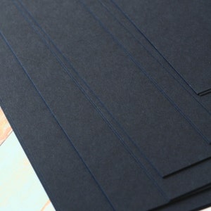May include: Stack of black cardstock paper. The paper has a textured surface and is arranged in a diagonal pattern.