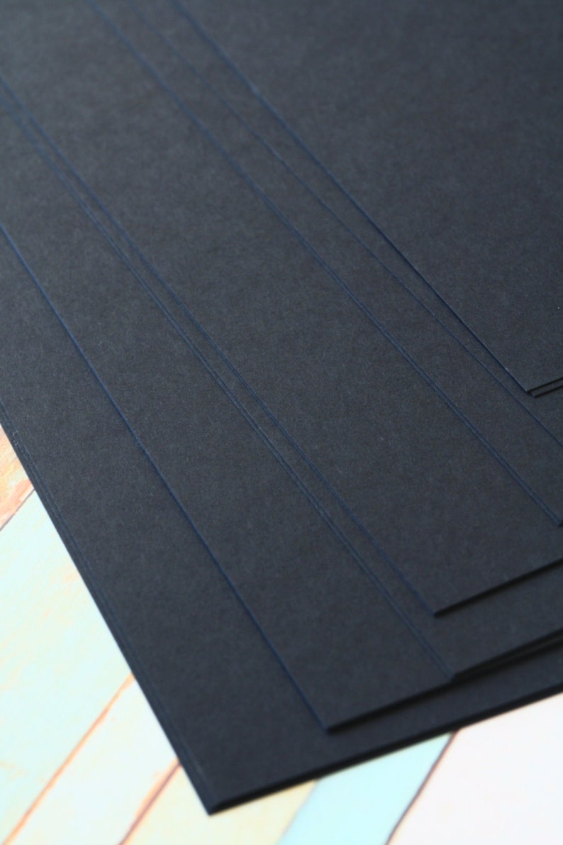 May include: Stack of black cardstock paper. The paper has a textured surface and is arranged in a diagonal pattern.