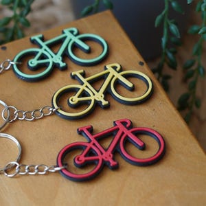 Puede incluir: Tres llaveros con forma de bicicleta en verde, amarillo y rojo. Cada llavero tiene una cadena y un anillo plateados. Los llaveros están sobre una superficie de madera, con vegetación en el fondo.