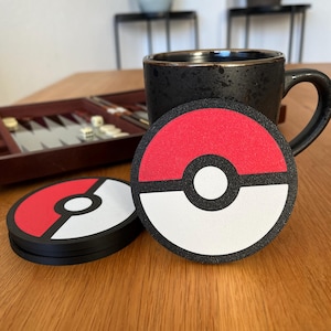 Könnte beinhalten: Ein Satz schwarzer, roter und weißer Untersetzer mit kreisförmigem Design. Die Untersetzer sind auf einer Holzoberfläche neben einer schwarzen Tasse mit Goldrand gestapelt. Das Design ähnelt einem Ball einer beliebten Zeichentrickfigur.