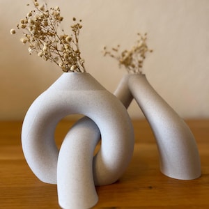 Puede incluir: Dos jarrones de cerámica gris claro con flores secas. Un jarrón tiene un diseño en bucle, y el otro tiene una forma arqueada y curvada. Los jarrones están sobre una superficie de madera, con un fondo neutro.