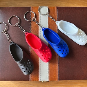 Puede incluir: Cuatro llaveros en miniatura con forma de zapato en negro, rojo, azul y blanco. Cada llavero presenta un pequeño dije de zapato detallado con una cadena y un llavero de color plateado. Los llaveros están colocados sobre una superficie a rayas marrón y blanca.